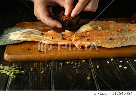 The cook uses his hands to add dry aromatic spices to raw fish fillets on the kitchen table. Concept of preparing fish herring in restaurant kitchen The cook uses his hands to add dry aromatic spices to raw fish fillets on the kitchen table. Concept of preparing fish herring in restaurant kitchen 112540454