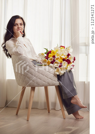Beautiful woman with spring flowers tulips in hands sitting on chair near window. Women's Day Beautiful woman with spring flowers tulips in hands sitting on chair near window. Women's Day 112541271