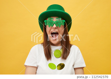 Saint Patrick's Day party. Extremely happy overjoyed woman wearing green hat and clover glasses standing isolated over yellow background screaming with excitement rejoicing Saint Patrick's Day party. Extremely happy overjoyed woman wearing green hat and clover glasses standing isolated over yellow background screaming with excitement rejoicing 112542370