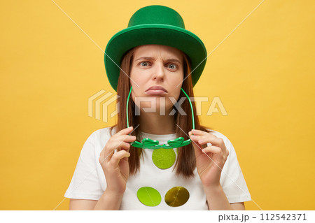 Irish holiday boring party. Sad upset unhappy woman wearing green hat holding clover glasses standing isolated over yellow background looking at camera with unhappy disappointed expression Irish holiday boring party. Sad upset unhappy woman wearing green hat holding clover glasses standing isolated over yellow background looking at camera with unhappy disappointed expression 112542371