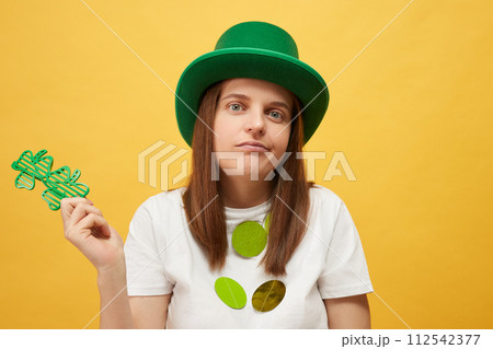 St. Patrick's day celebration Uncertain woman wearing green leprechaun hat and shamrock glasses posing isolated over yellow background shrugging shoulders 112542377