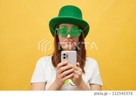 Celebrating St. Patrick's Day traditions. Disappointed woman wearing green leprechaun hat and shamrock glasses posing isolated over yellow background using mobile phone with sad face 112542378