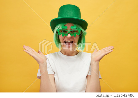 Funny woman with green hair wearing leprechaun hat and shamrock glasses posing isolated over yellow background spreading her face exclaiming with excitement Funny woman with green hair wearing leprechaun hat and shamrock glasses posing isolated over yellow background spreading her face exclaiming with excitement 112542400