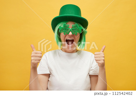 Overjoyed excited woman with green hair wearing leprechaun hat and shamrock glasses posing isolated over yellow background showing like gesture thumbs up Overjoyed excited woman with green hair wearing leprechaun hat and shamrock glasses posing isolated over yellow background showing like gesture thumbs up 112542402