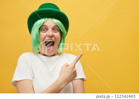 Amazed excited woman with green hair wearing leprechaun hat posing isolated over yellow background pointing at copy space for promotional text indicating at advertisement area Amazed excited woman with green hair wearing leprechaun hat posing isolated over yellow background pointing at copy space for promotional text indicating at advertisement area 112542403