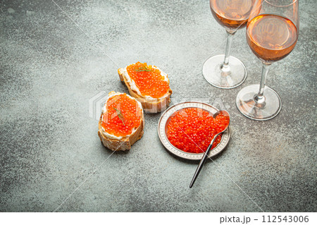 Small metal plate with red salmon caviar, two caviar toasts canape, two glasses of champagne top view on grey concrete background, festive luxury delicacy and appetizer Small metal plate with red salmon caviar, two caviar toasts canape, two glasses of champagne top view on grey concrete background, festive luxury delicacy and appetizer 112543006