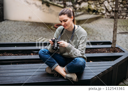 Young tourist woman smiling and using vintage camera at the city. Happy woman holding her chin and looking at the camera while sitting on bench outdoors, traveler 112543361