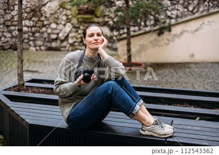 Young tourist woman smiling and using vintage camera at the city. Happy woman holding her chin and looking at the camera while sitting on bench outdoors, traveler 112543362
