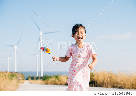 A joyful child holding pinwheels runs near windmills embodying wind energy education. Clean electricity and sustainable industry visuals in a picturesque wind turbine landscape. 112546940