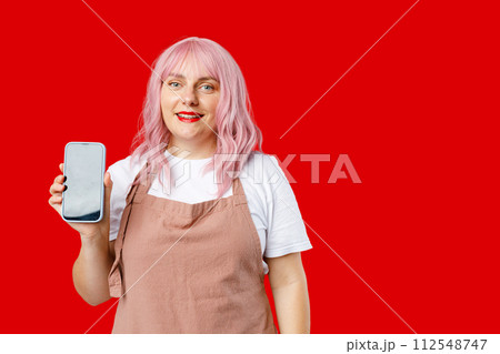 Great Mobile Offer. Excited happy 30s woman with pink hair in apron holding Smartphone in her hand, Emotionally Reacting To New App. Female posing on red background, studio portrait. 112548747