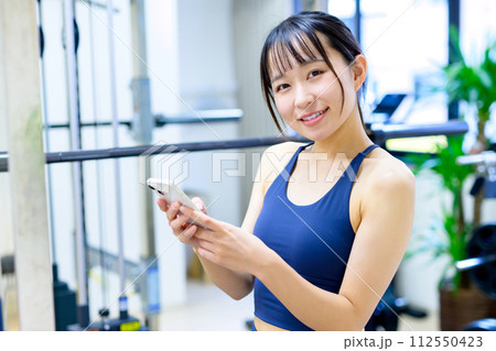 ジムでスマホを見る女性　SNS　体調管理アプリ　入会　予約　計画　フィットネス　トリミングアップ 112550423