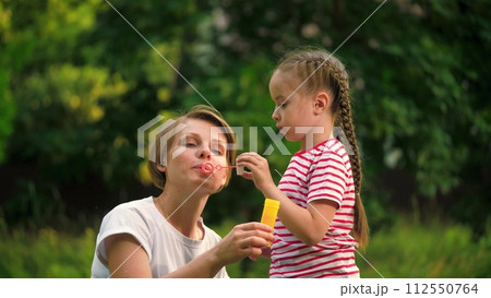 Loving mother with cheerful daughter blows bubbles in park among trees on vacation. Mother waits till preschooler girl blows bubbles in meadow on weekend. Mother and child spend time blowing bubbles Loving mother with cheerful daughter blows bubbles in park among trees on vacation. Mother waits till preschooler girl blows bubbles in meadow on weekend. Mother and child spend time blowing bubbles 112550764