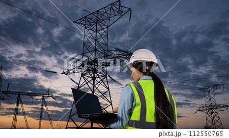 Woman electrician marks notes online on laptop observing power transmission lines at twilight countryside closeup. Engineer tests system with laptop at power generation plant under dramatic sky 112550765