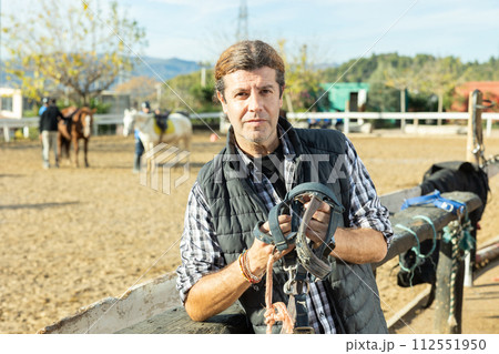 Portrait of skilled adult male stable worker holding horse halter or harness near wooden fence on ranch 112551950