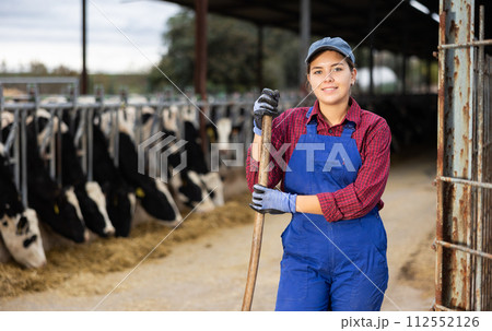 Portrait of smiling European female farmer in uniform with rake during work on dairy farm Portrait of smiling European female farmer in uniform with rake during work on dairy farm 112552126