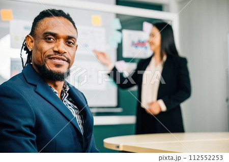 In an office meeting, a black businessman, a charismatic CEO and leader, smiles at the camera. His presence exudes confidence and success. A portrait of leadership and diversity in a corporate setting 112552253