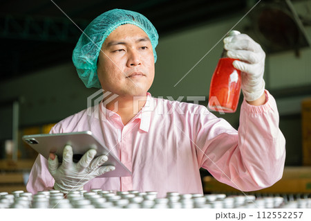 Quality control worker inspects bottles on conveyor belt in beverage factory ensuring top industry standards. Engineer and inspector collaborate on tablet for efficient production. 112552257