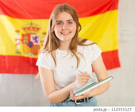 Portrait of a smiling satisfied girl student with notebooks in her hands against the background of flag of Spain Portrait of a smiling satisfied girl student with notebooks in her hands against the background of flag of Spain 112553127