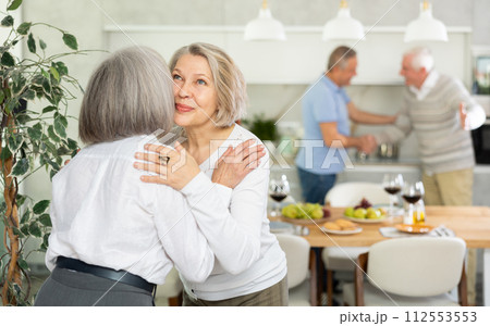 Two elderly woman greet each other by kissing each other on the cheek. While men shake hands Two elderly woman greet each other by kissing each other on the cheek. While men shake hands 112553553