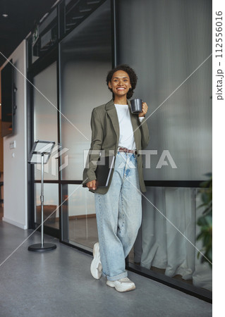 A woman in formal wear holds a coffee cup and laptop in a hallway A woman in formal wear holds a coffee cup and laptop in a hallway 112556016