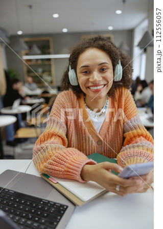 A woman with headphones sits at a table with laptop and cell phone in a cozy minimalistic cafe 112556057