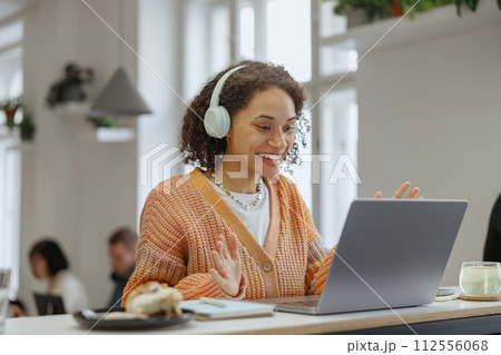 A woman with headphones at a table using a laptop and smiling in a cozy minimalistic cafe 112556068