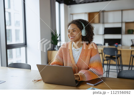 A woman with headphones using a laptop at a wooden table in nice modern coworking office 112556092