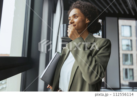 A woman in formal wear is gazing out a window, laptop in hand, in nice modern coworking office 112556114