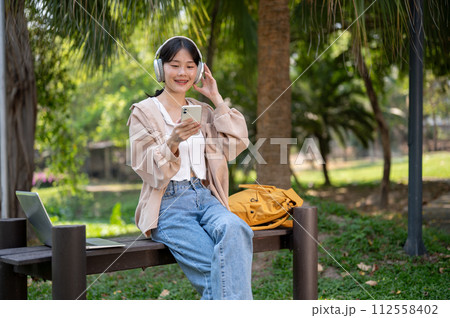 A pretty Asian female student is relaxing on a bench in a park, listening to music on her headphones A pretty Asian female student is relaxing on a bench in a park, listening to music on her headphones 112558402