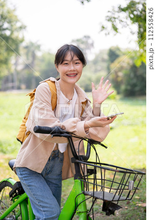 A cheerful Asian female college student waves her hand, greeting her friends while riding her bike. A cheerful Asian female college student waves her hand, greeting her friends while riding her bike. 112558426