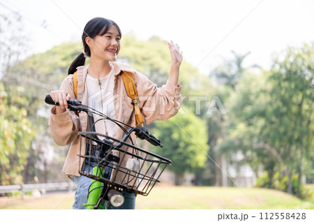 A cheerful Asian female college student waves her hand, greeting her friends while riding her bike. A cheerful Asian female college student waves her hand, greeting her friends while riding her bike. 112558428