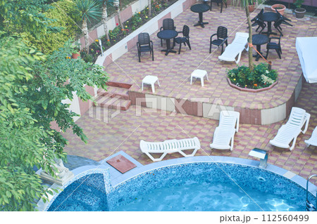 Top view of the hotel territory with round outdoor swimming pool with blue mosaic ceramic tiles, white plastic sun loungers, tables and chairs, trees and palm trees. Background with copy space. 112560499