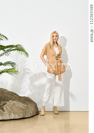 Trendy blonde woman in sleeveless top and loose trousers posing under hard sunlight near the rock 112563389
