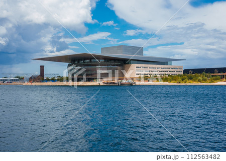 A modern, layered architectural marvel reflects the sky on its glass and metal facade, likely the Copenhagen Opera House, set against a calm waterfront under a partly cloudy sky. 112563482