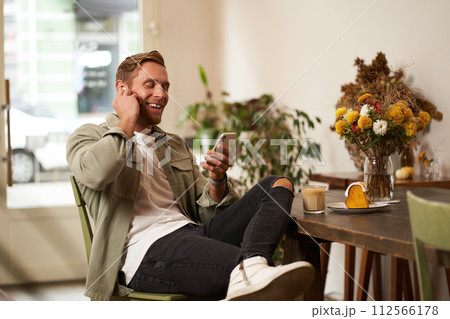 Lifestyle shot of handsome young man, looking at his mobile phone, listening to music in wireless earphones, sitting in a cafe, drinking coffee 112566178