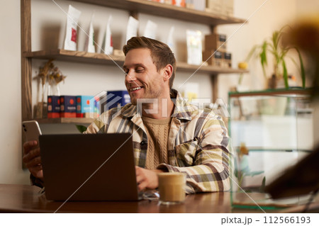 Lifestyle portrait of young businessman, freelancer sitting in cafe, drinking coffee and using laptop, working on online project, digital nomad doing his task, has remote job, looking satisfied 112566193