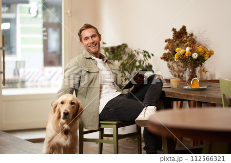Portrait of handsome young man, cafe visitor, sitting with his dog, petting golden retriever and reading news on digital tablet, concept of lifestyle and pet-friendly public places Portrait of handsome young man, cafe visitor, sitting with his dog, petting golden retriever and reading news on digital tablet, concept of lifestyle and pet-friendly public places 112566321