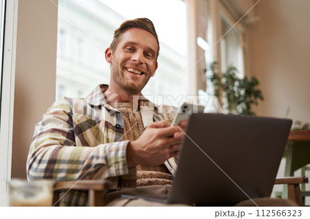 Portrait of smiling, handsome young man relaxing in coffee shop, working remotely from cafe co-working space, using laptop and smartphone, looking at camera Portrait of smiling, handsome young man relaxing in coffee shop, working remotely from cafe co-working space, using laptop and smartphone, looking at camera 112566323