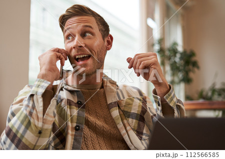Freelance, digital nomad and workspace concept. Portrait of handsome young man with mobile phone, sitting in cafe with laptop, working on project, studying remotely, looking back outside window 112566585