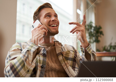 Lifestyle shot of young happy man sitting in cafe with laptop, answer phone call, talking to friend on mobile and laughing, chatting to someone with joyful face, resting in chair 112566668