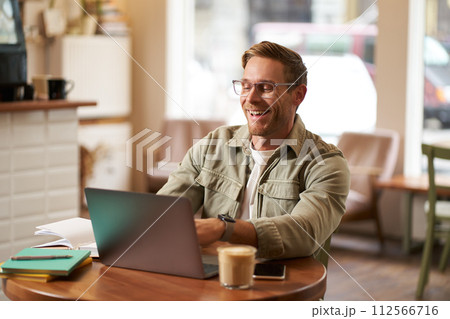 Image of young digital nomad, man in glasses sits in cafe, works from coffee shop, uses laptop in co-working space, wears glasses, drinks his beverage 112566716
