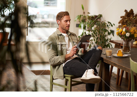 Lifestyle shot of handsome young man sitting in a cafe in front of the table, drinking coffee, wearing wireless headphones, listening to music or watching video in public space, using digital tablet 112566722