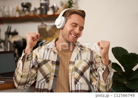 Portrait of handsome happy guy, freelancer working and listening to music in wireless headphones, dancing on his chair in cafe, enjoying favourite song in earphones Portrait of handsome happy guy, freelancer working and listening to music in wireless headphones, dancing on his chair in cafe, enjoying favourite song in earphones 112566749