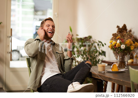 Lifestyle portrait of handsome young man, sitting in cafe, talking to a friend on mobile phone with wireless earphones, laughing and smiling, listening to funny podcast 112566920