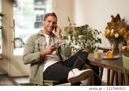 Lifestyle portrait of handsome stylish young man, sits in cafe, holds mobile phone, puts on wireless earphones, looks at camera with happy smile Lifestyle portrait of handsome stylish young man, sits in cafe, holds mobile phone, puts on wireless earphones, looks at camera with happy smile 112566932