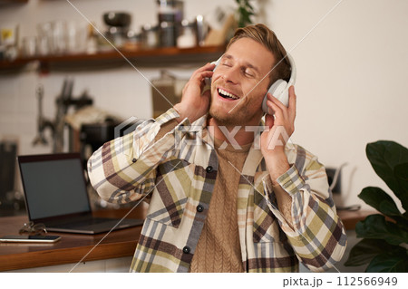 Handsome smiling young man, digital nomad, sitting in co-working open space, working in coffee shop, singing and listening to music in wireless headphones Handsome smiling young man, digital nomad, sitting in co-working open space, working in coffee shop, singing and listening to music in wireless headphones 112566949