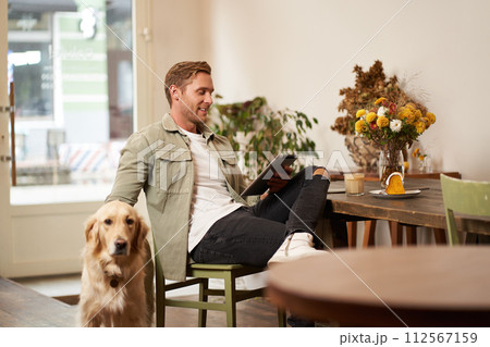 Portrait of handsome young man sits in cafe with his dog, petting golden retriever, reading news on tablet, relaxing with cup of coffee Portrait of handsome young man sits in cafe with his dog, petting golden retriever, reading news on tablet, relaxing with cup of coffee 112567159