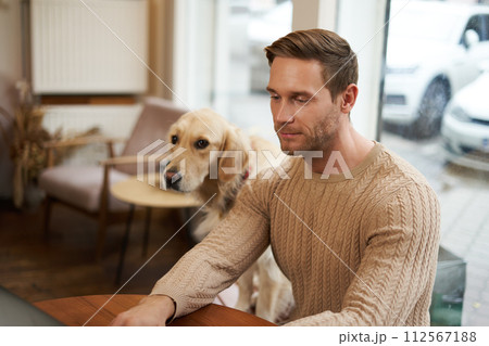 Handsome young man, digital nomad sitting with his dog in a pet-friendly cafe, working on laptop while a golden retriever watches him Handsome young man, digital nomad sitting with his dog in a pet-friendly cafe, working on laptop while a golden retriever watches him 112567188
