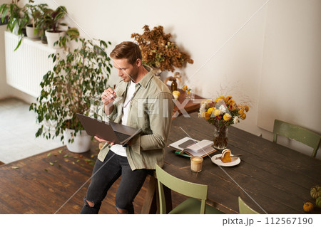 Portrait of young manager, man sits on table, holds laptop, looks concentrated at screen, reading project information, checking work data, working in office or co-working space 112567190
