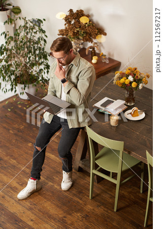 Vertical shot of handsome stylish businessman, standing near the table with laptop, thinking, working on a project on his computer, studying online, doing freelance task 112567217